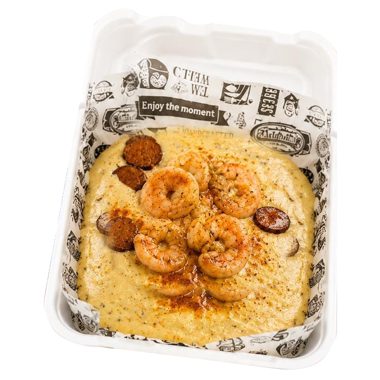 Shrimp and grits topped with sausage slices in a takeout container